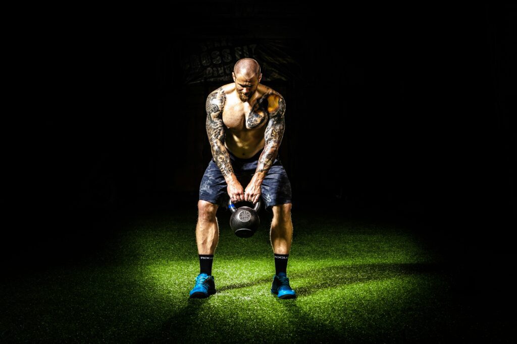 Muscular man with tattoos lifting a kettlebell indoors under a spotlight. Intense fitness and strength training.