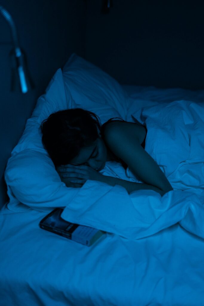Woman sleeping peacefully in cozy blue-lit bedroom with book beside her.