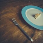Minimalist bread piece on a blue-rimmed plate with fork and knife on wooden table.