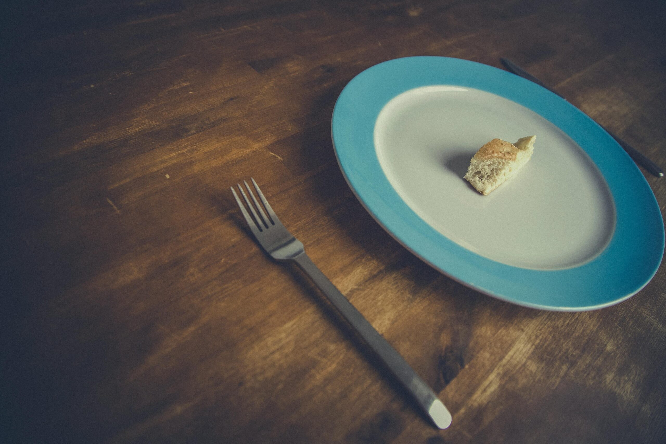 Minimalist bread piece on a blue-rimmed plate with fork and knife on wooden table.