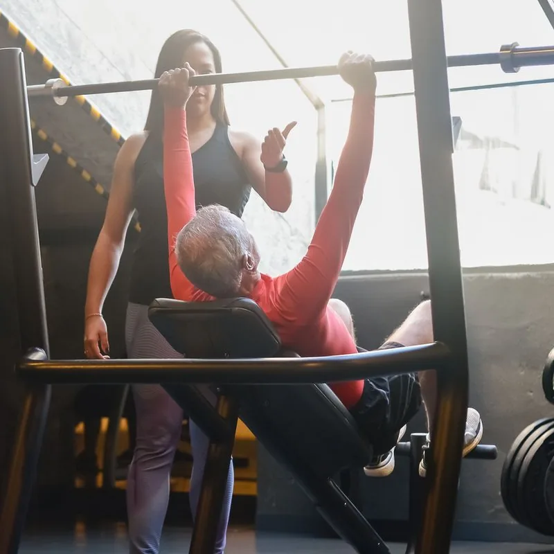 Homem mais velho realizando supino com acompanhamento de uma treinadora